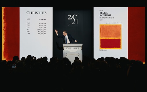 An auctioneer stands onstage at Christie’s, announcing the sale of Mark Rothko’s No. 31 (Yellow Stripe) painting to a full audience.