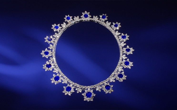 A diamond and sapphire necklace, elegantly designed with floral motifs, is showcased against a deep blue background. | An elegant, diamond and sapphire tiara displayed against a deep blue backdrop.