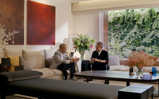 Two individuals converse in a stylish living room adorned with lush flowers and large windows revealing a verdant outdoor view.