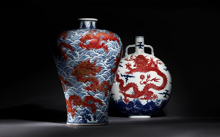 Two ornate vases with red dragons and blue waves are displayed against a dark background. | Two ornate Chinese porcelain vases feature red dragons and blue waves on a dark background.