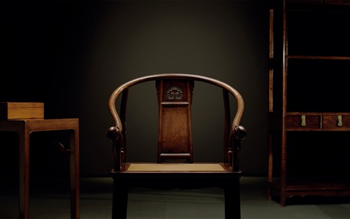 A classic wooden armchair is centered between a table and a cabinet against a dark background.