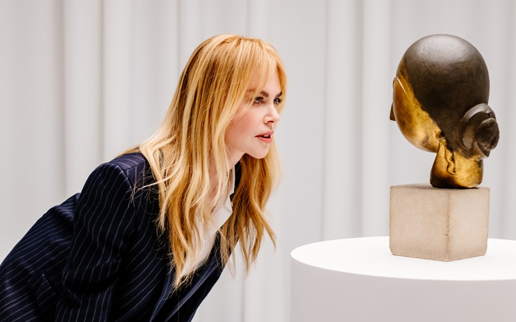 A person with long blonde hair observes a sculpted head displayed on a pedestal.