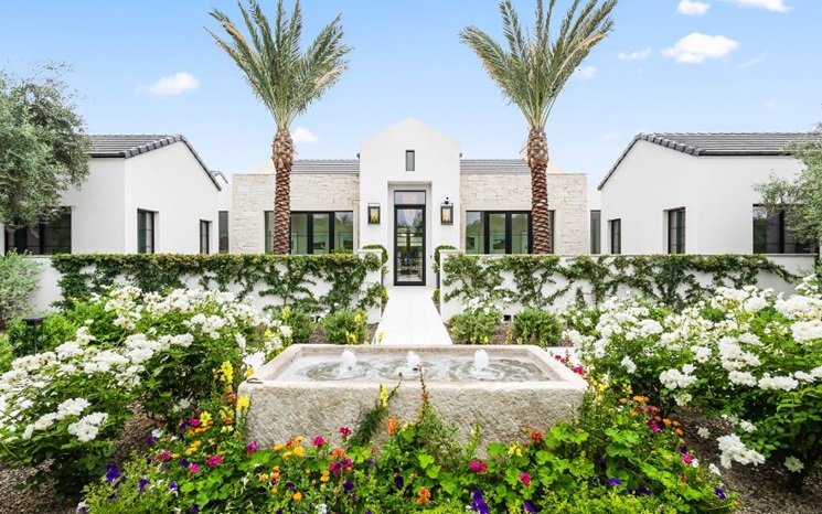 A modern white house with palm trees, a colorful garden, and a stone fountain in front.