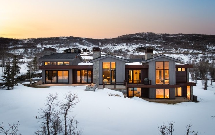 Modern house with large windows surrounded by snowy landscape at sunset.