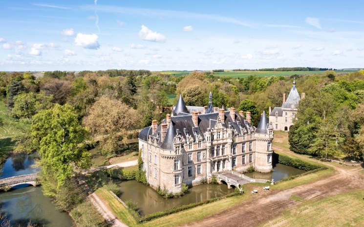 A large, ornate castle sits surrounded by a moat and lush greenery under a blue sky.