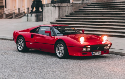 A classic red sports car with pop-up headlights illuminated, parked near stairs.