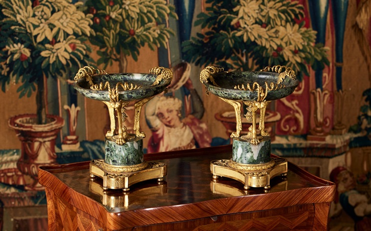 Two ornate, green marble and gold decorative bowls sit on a polished wooden table against a tapestry.