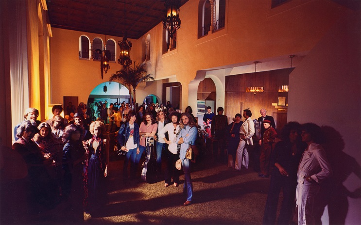 A large group of people is gathered in a warmly lit, spacious room with high ceilings and arches.