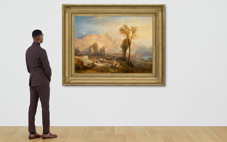 A man dressed in a suit admires a classic landscape painting featuring a tree and distant mountains in an art gallery.