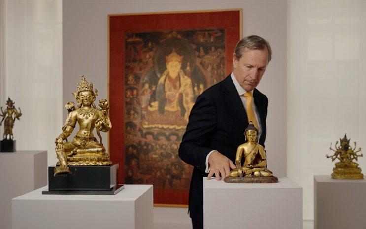 The image shows an art gallery with various golden Buddhist statues and intricate tapestries displayed on pedestals.