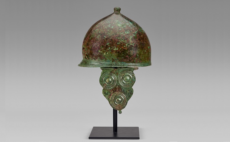 An ancient bronze helmet with a domed top and decorative cheek guards on display.