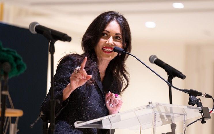 A woman is speaking at a podium with microphones, gesturing with her hand and smiling.