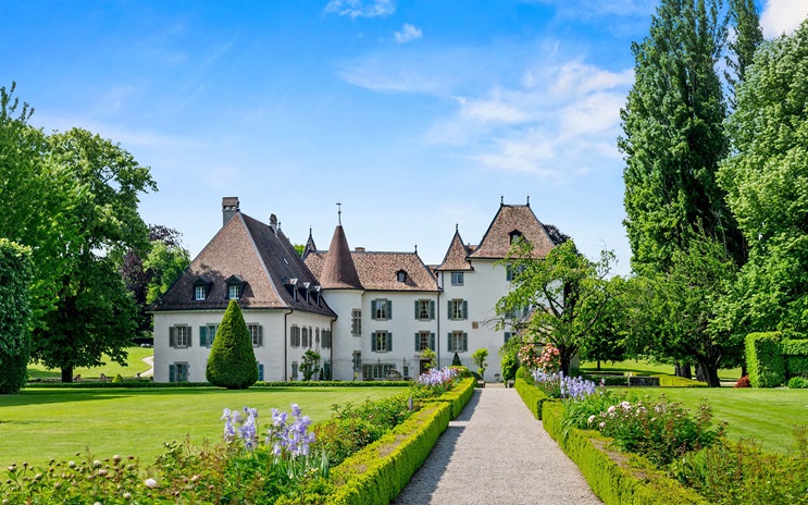 A large white mansion with a pointed roof sits surrounded by gardens and trees under a blue sky.