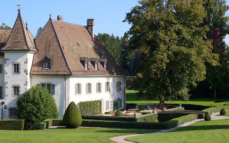 A large house with a steep tiled roof sits beside a manicured garden with hedges and trees.
