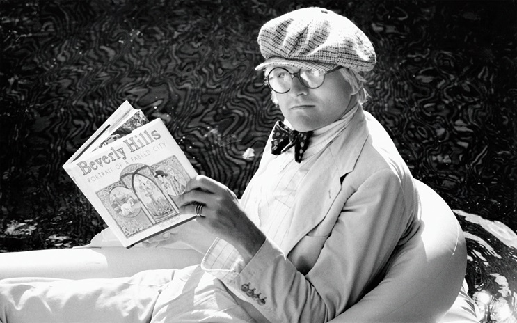 David Hockney in a light suit and hat reads a "Beverly Hills" book while floating on a pool raft.