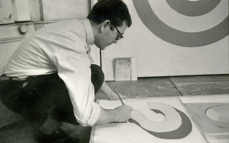 Joe Tilson is painting abstract circular shapes on canvas while crouching on the floor.