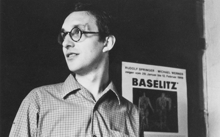 Georg Baselitz in a checkered shirt stands beside a poster with the text "BASELITZ" on it.