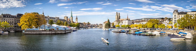 Panoramic view of Zurich with historic buildings, a river filled with boats, and tree-lined promenades under a clear sky.