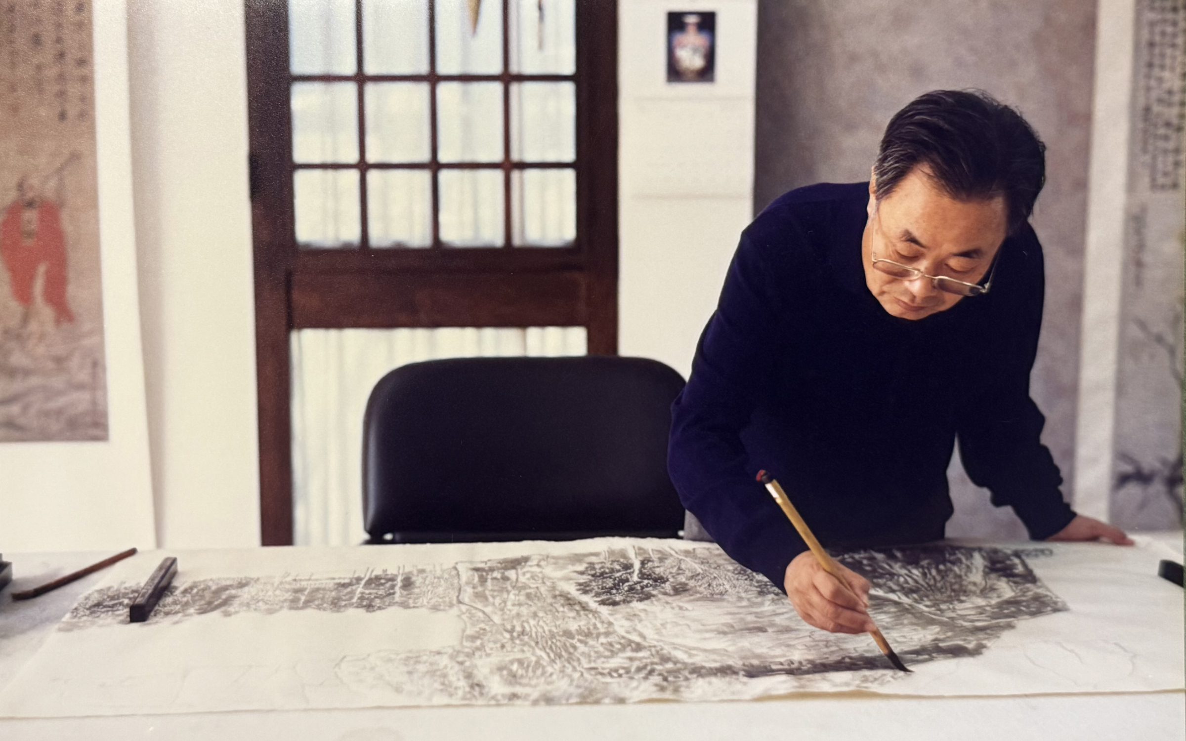 A person is drawing on a large paper on a table in a room with traditional decor and artwork hanging on the walls.