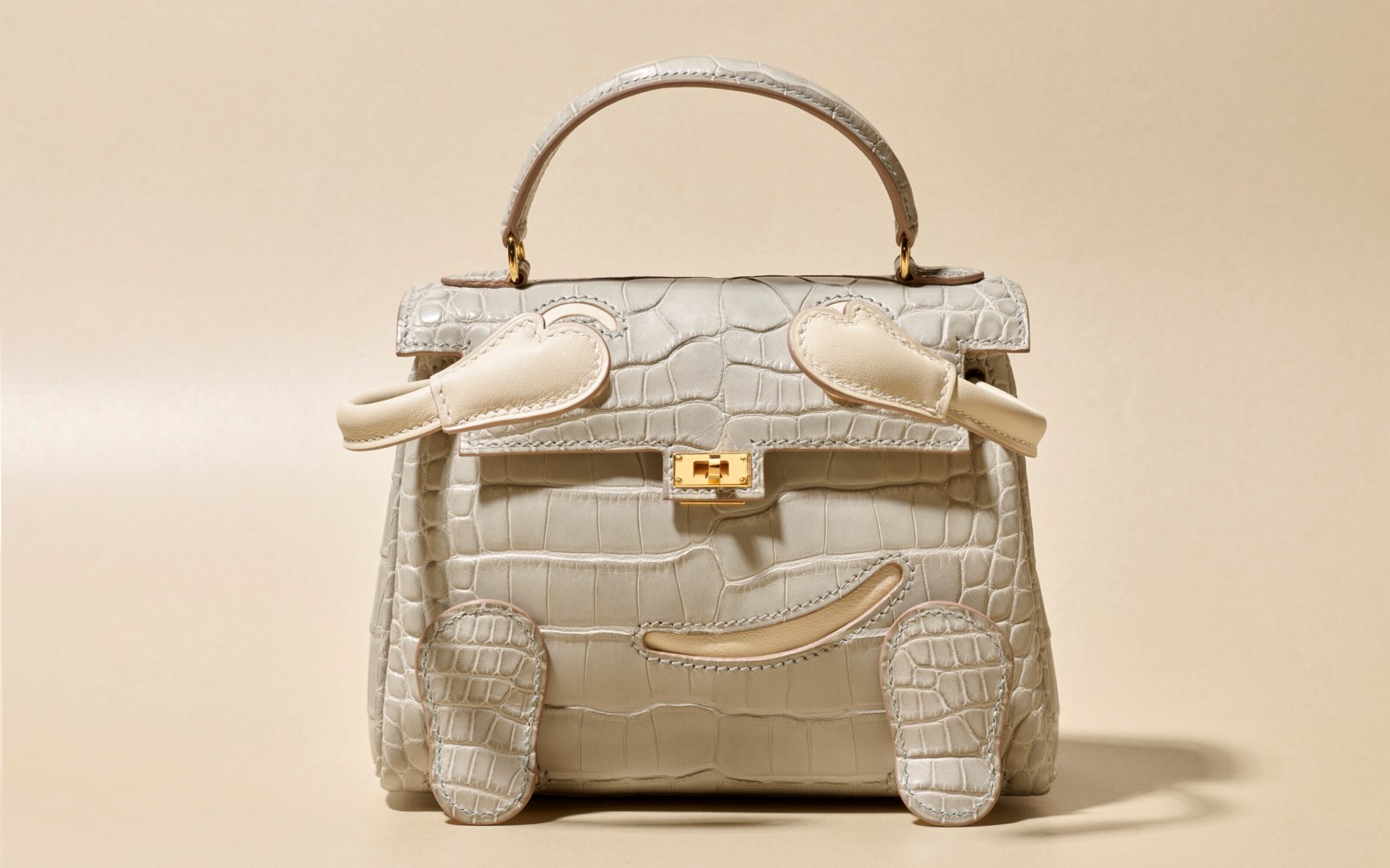 A light gray, crocodile leather handbag with intricate detailing and a metallic clasp, displayed against a neutral background.