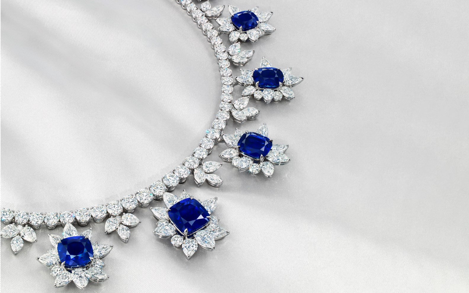 An elegant necklace with large blue gems surrounded by intricate diamond patterns on a light gray background.
